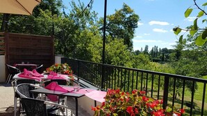 Une terrasse extérieure avec des tables et des chaises, entourée d’une rambarde en métal, et un grand pot de fleurs contenant des plantes aux fleurs rouges.