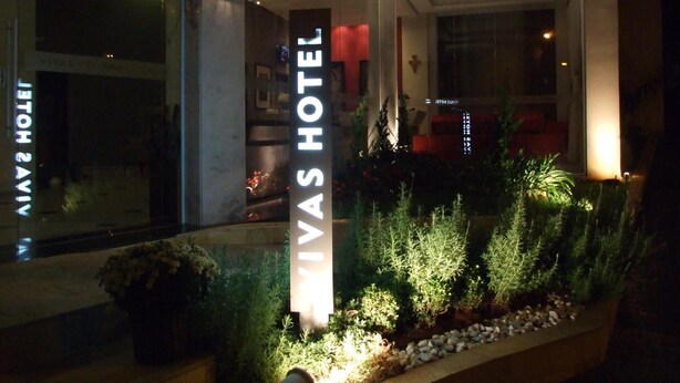 Exterior do Hotel Vivas, com uma placa em destaque, entrada bem iluminada e área de jardim ajardinada.