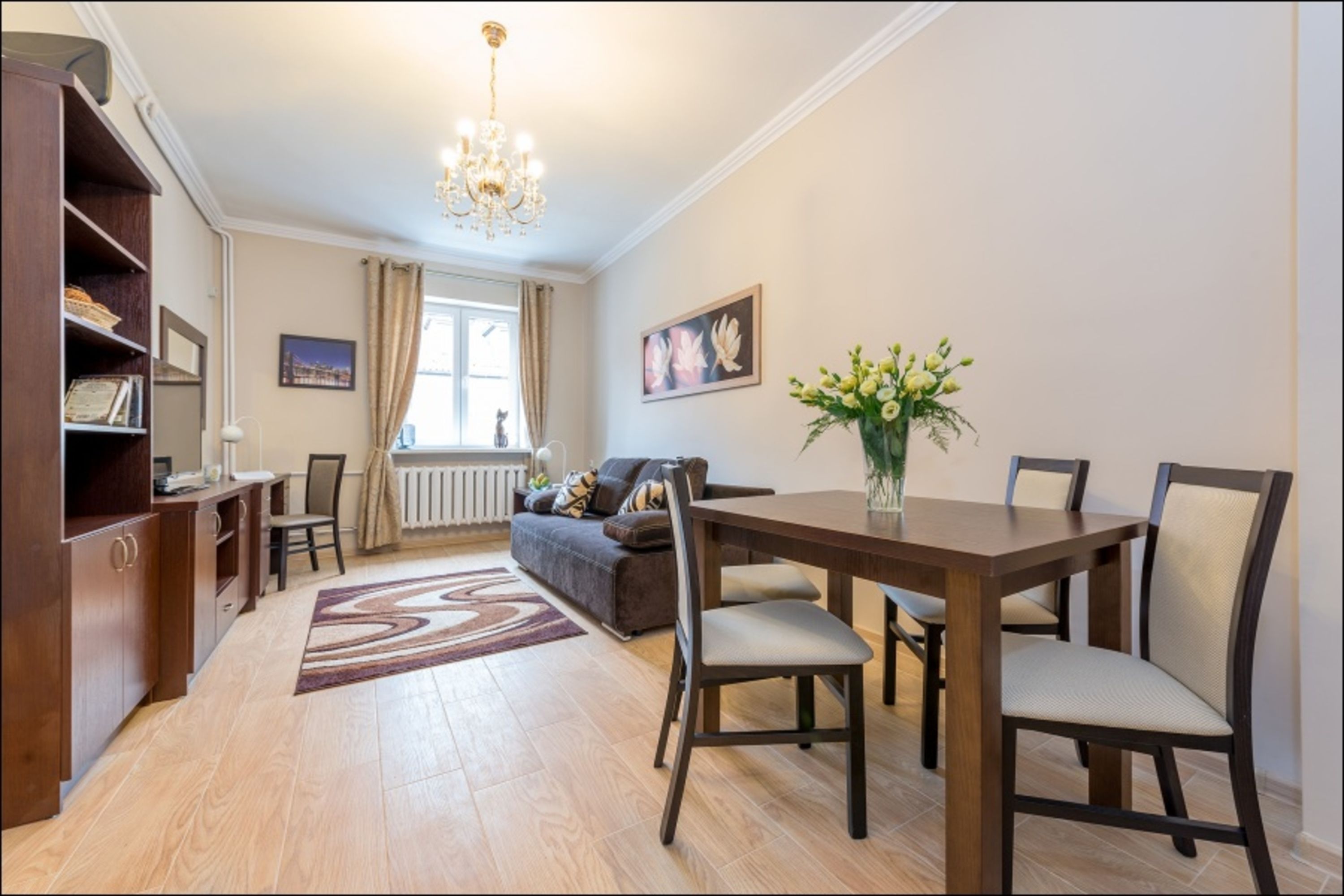 A dining area with a wooden table and chairs, a chandelier, a sofa, and a radiator.