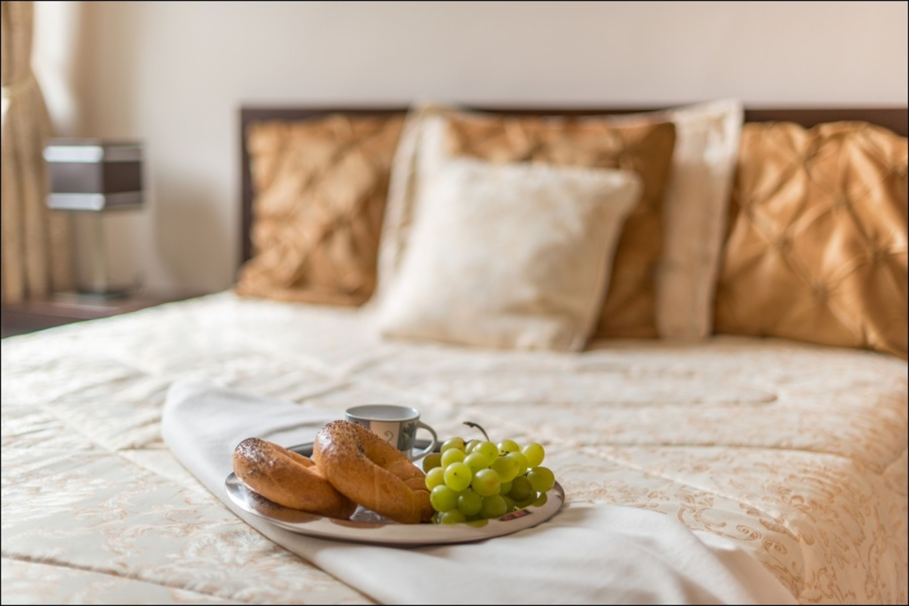 Una cama bien hecha con un plato de croissants y uvas, una taza de café y una mesita de noche con una lámpara.