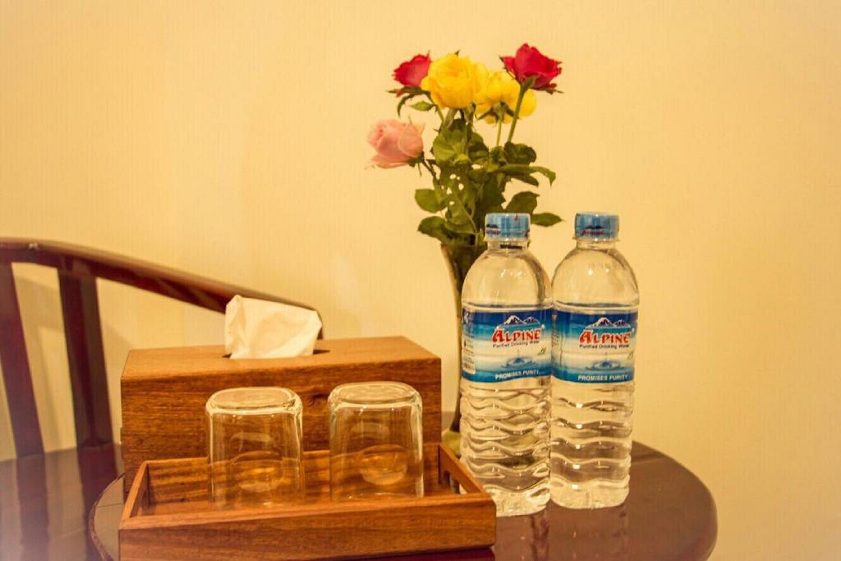 A wooden tray with three glasses, two water bottles, and a vase with flowers on a table.