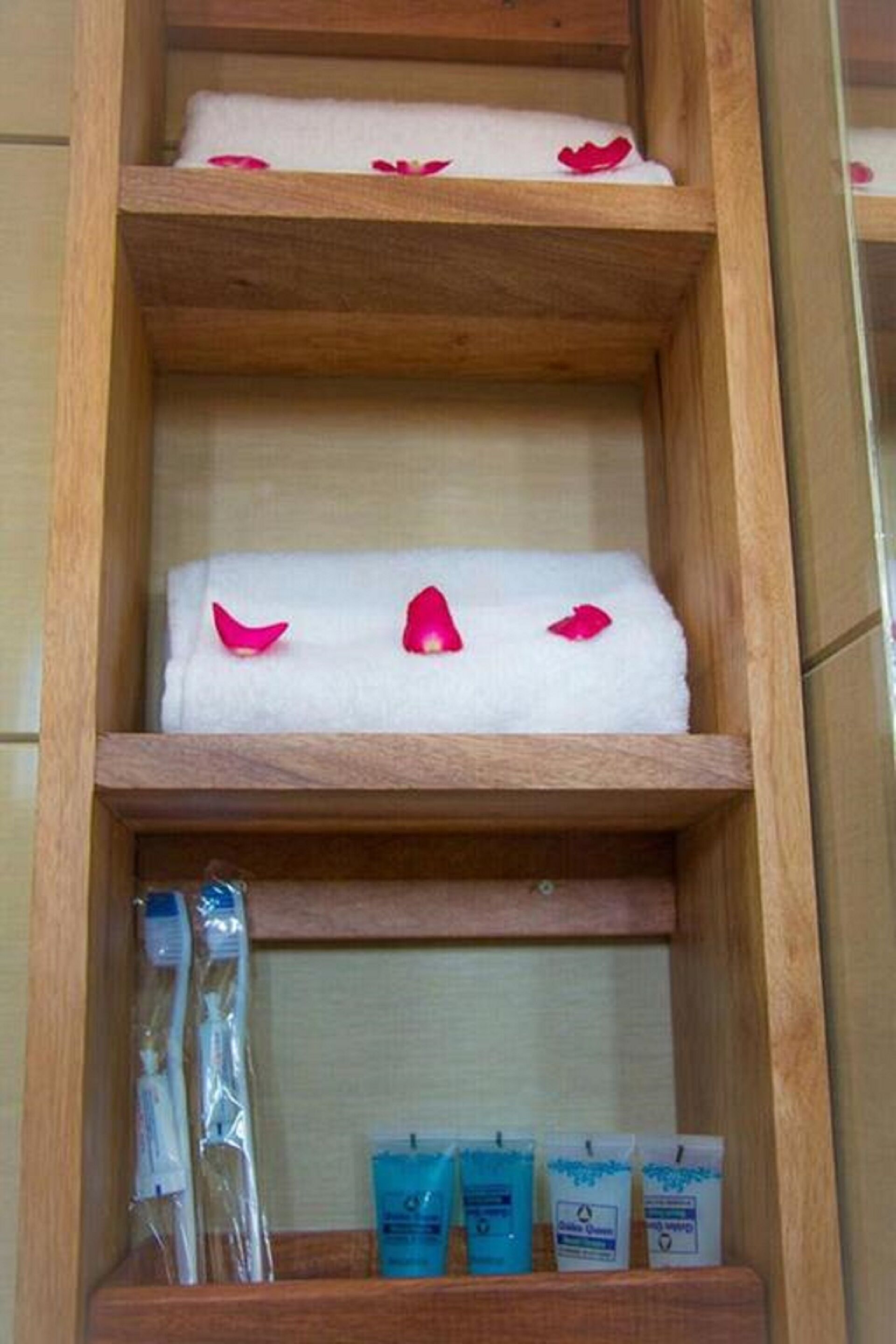 A wooden shelf with a white towel and rose petals, and three blue toiletry packets.
