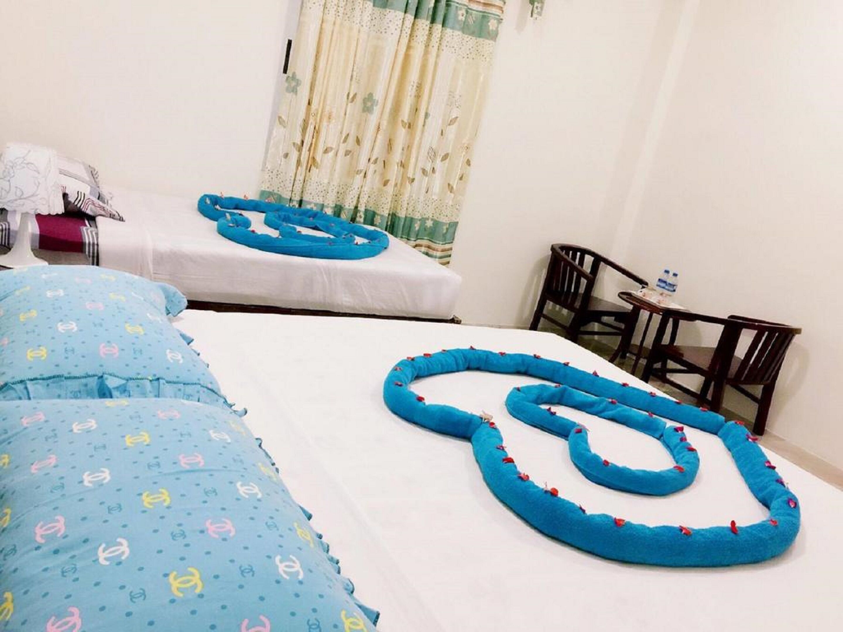 A hotel room with two beds, each with blue bedding and decorative pillows. There is a small table with chairs and a water bottle on it.