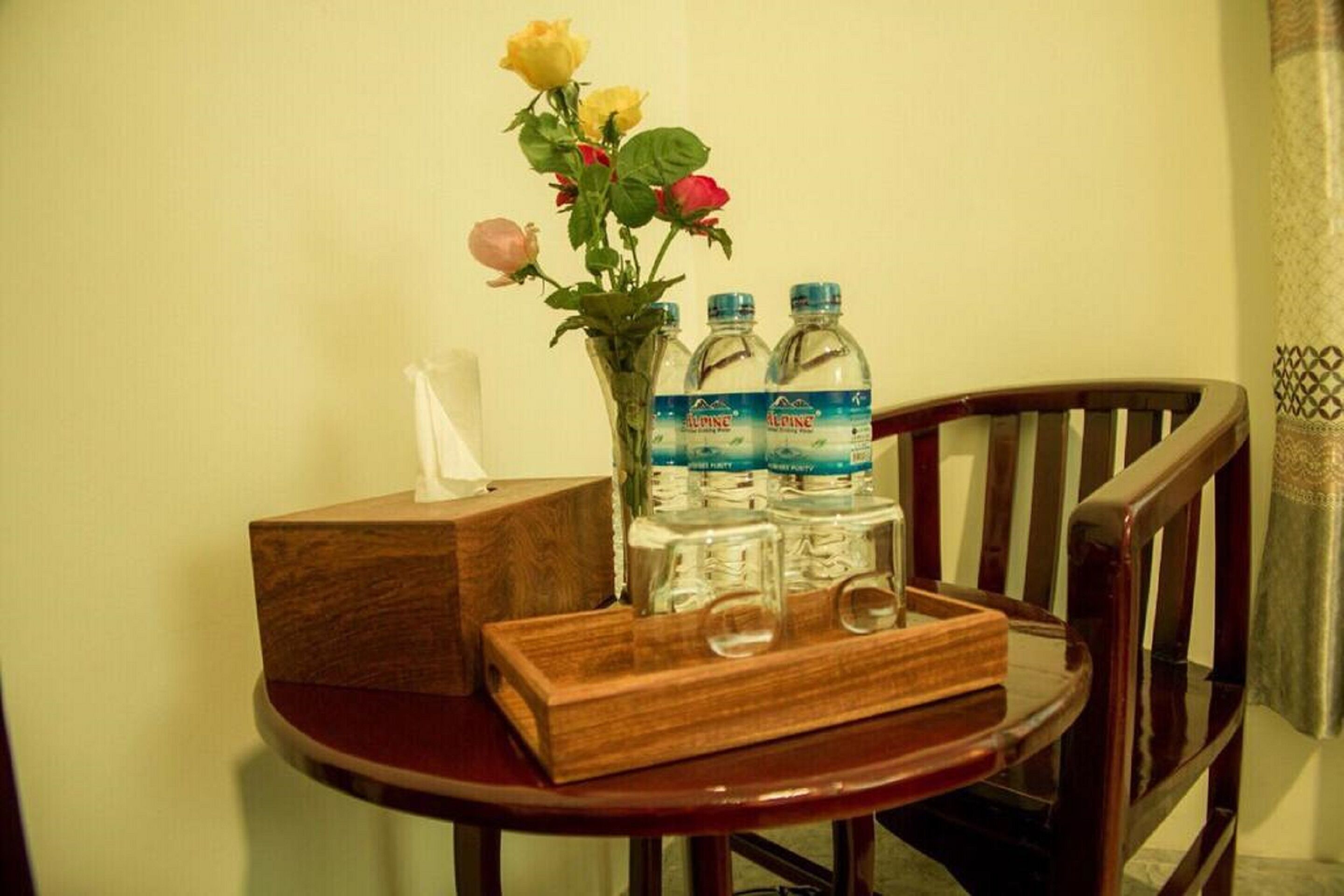 Uma mesa de madeira com uma caixa de lenços, um vaso com rosas e três garrafas de água.