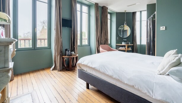 Ein Schlafzimmer mit einem großen Bett, einem kleinen Tisch, einem Stuhl, einem Spiegel und einem Fenster mit Vorhängen.