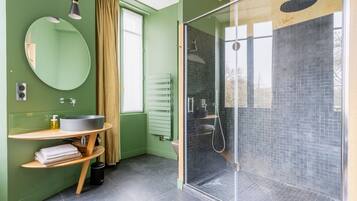 A modern bathroom with a green wall, a round mirror, a wooden vanity, a glass-enclosed shower, and a window with curtains.