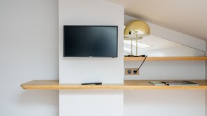 Ein modernes Zimmer mit montiertem Fernseher, einem Schreibtisch inklusive Fernbedienung und einer Lampe.