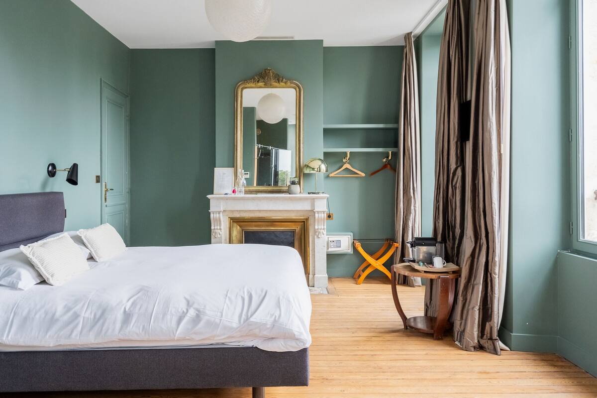 Un dormitorio con cama, chimenea, espejo y ventana con cortinas.