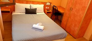 A neatly made bed with a grey quilt, a black pillow, and a rolled white towel. There is a wooden nightstand with a lamp and a small table with a chair. - Hotel Restaurant Les Alizes (Le Pouzin)
