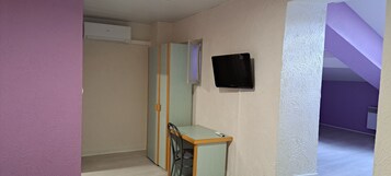 Una habitación con cama, mesita de noche, televisor montado en la pared y una puerta que da a otra habitación.