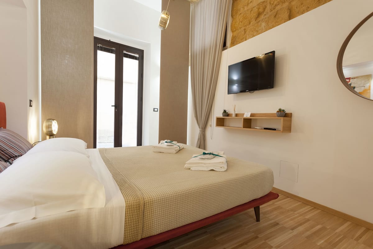 Habitación de hotel con cama, televisión montada en la pared, estante de madera con planta y un espejo grande.
