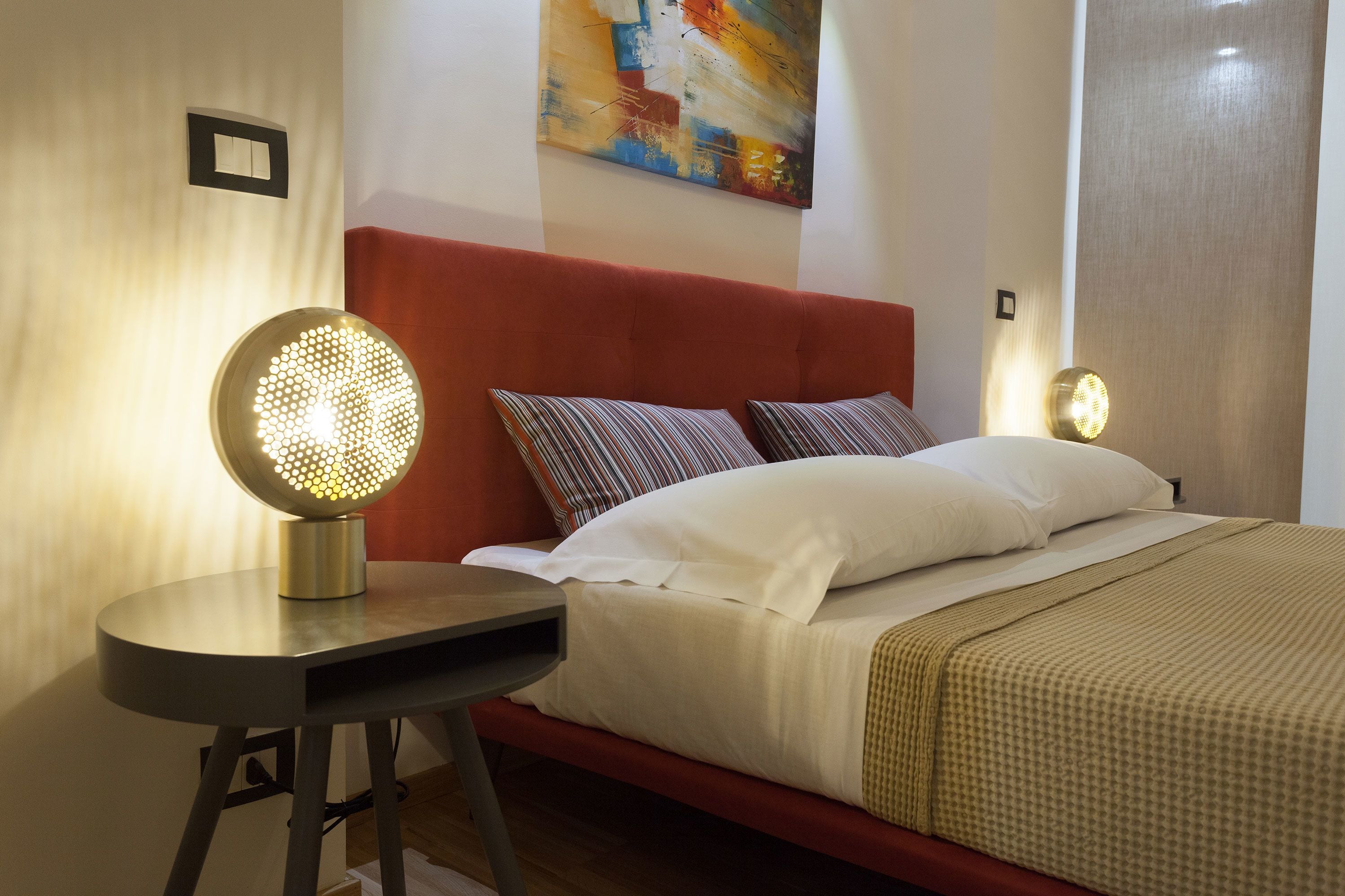 Un lit bien fait, agrémenté d’oreillers blancs et à motifs, d’une tête de lit rouge et d’une table ronde surmontée d’un luminaire.