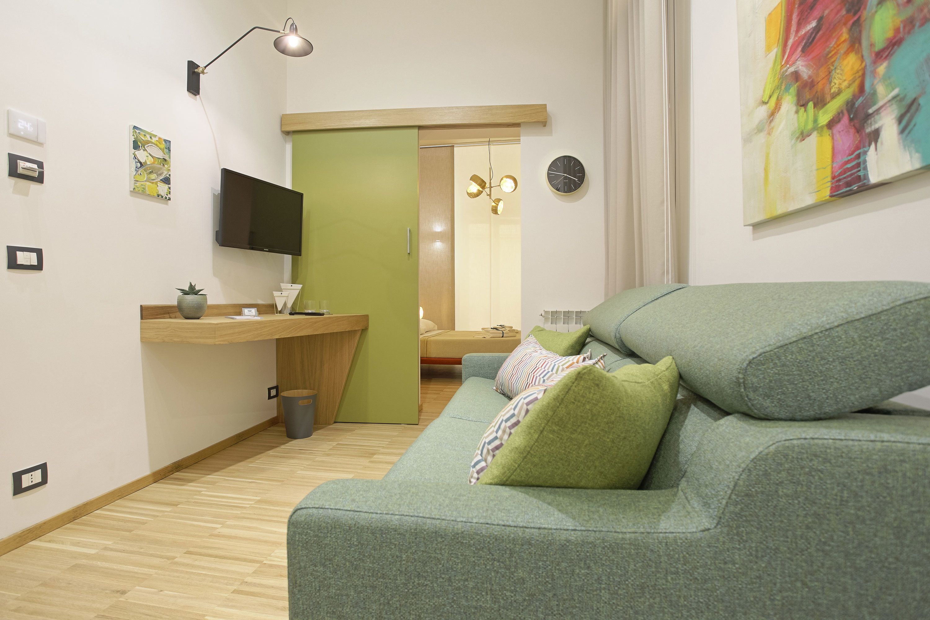 Un salon moderne avec un canapé vert, un bureau en bois, un téléviseur à écran plat et un tableau abstrait aux couleurs vives.
