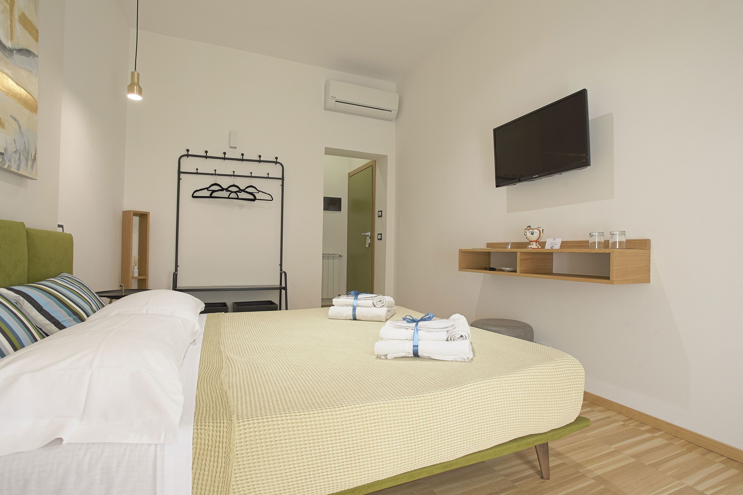 Habitación de hotel con una cama, un televisor montado en la pared, un perchero y un estante pequeño con artículos de aseo.