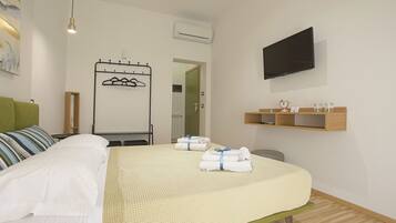 Habitación de hotel con una cama, un televisor montado en la pared, un perchero y un estante pequeño con artículos de aseo.