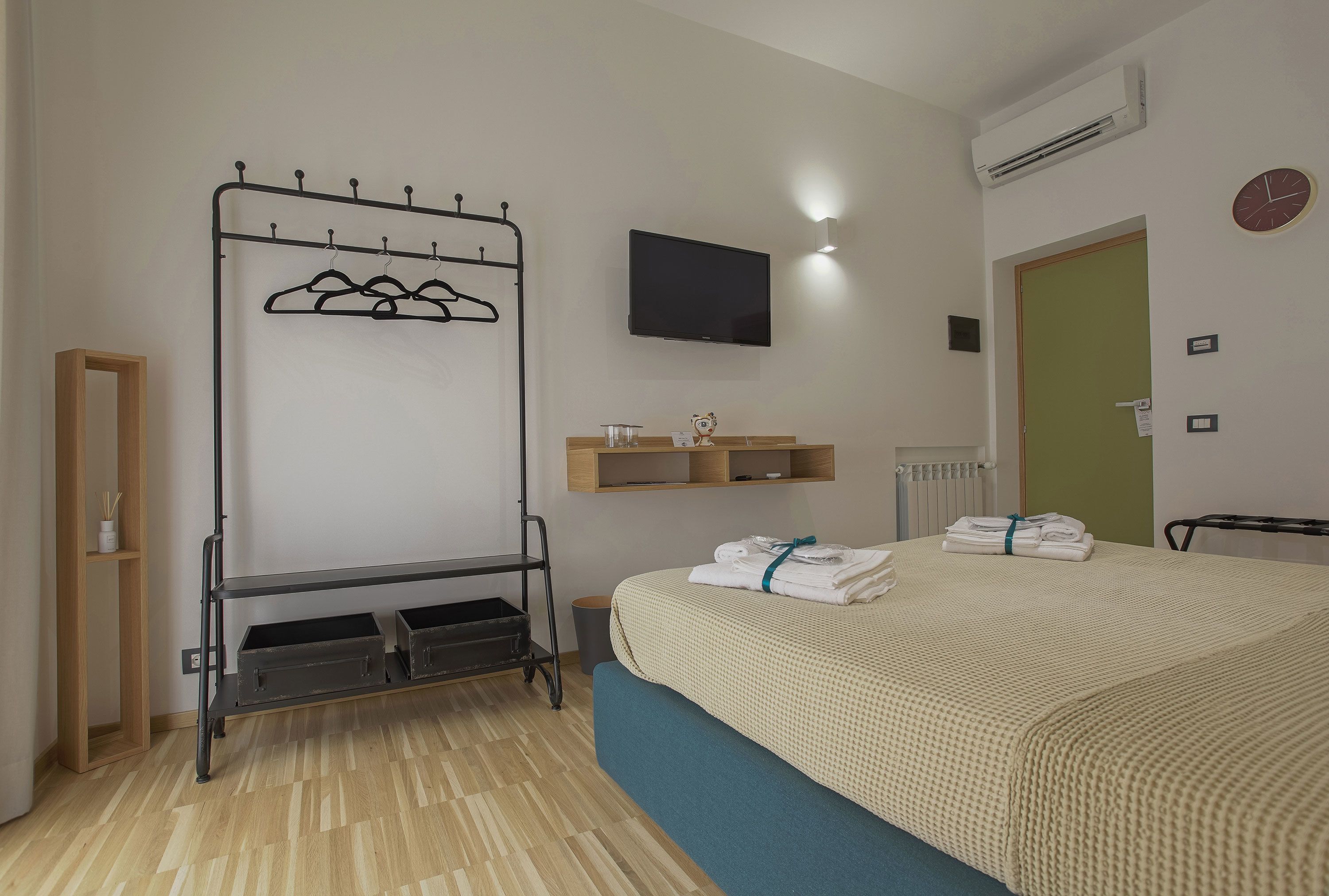 Habitación de hotel con cama, televisor, perchero, repisa con artículos de aseo y radiador.