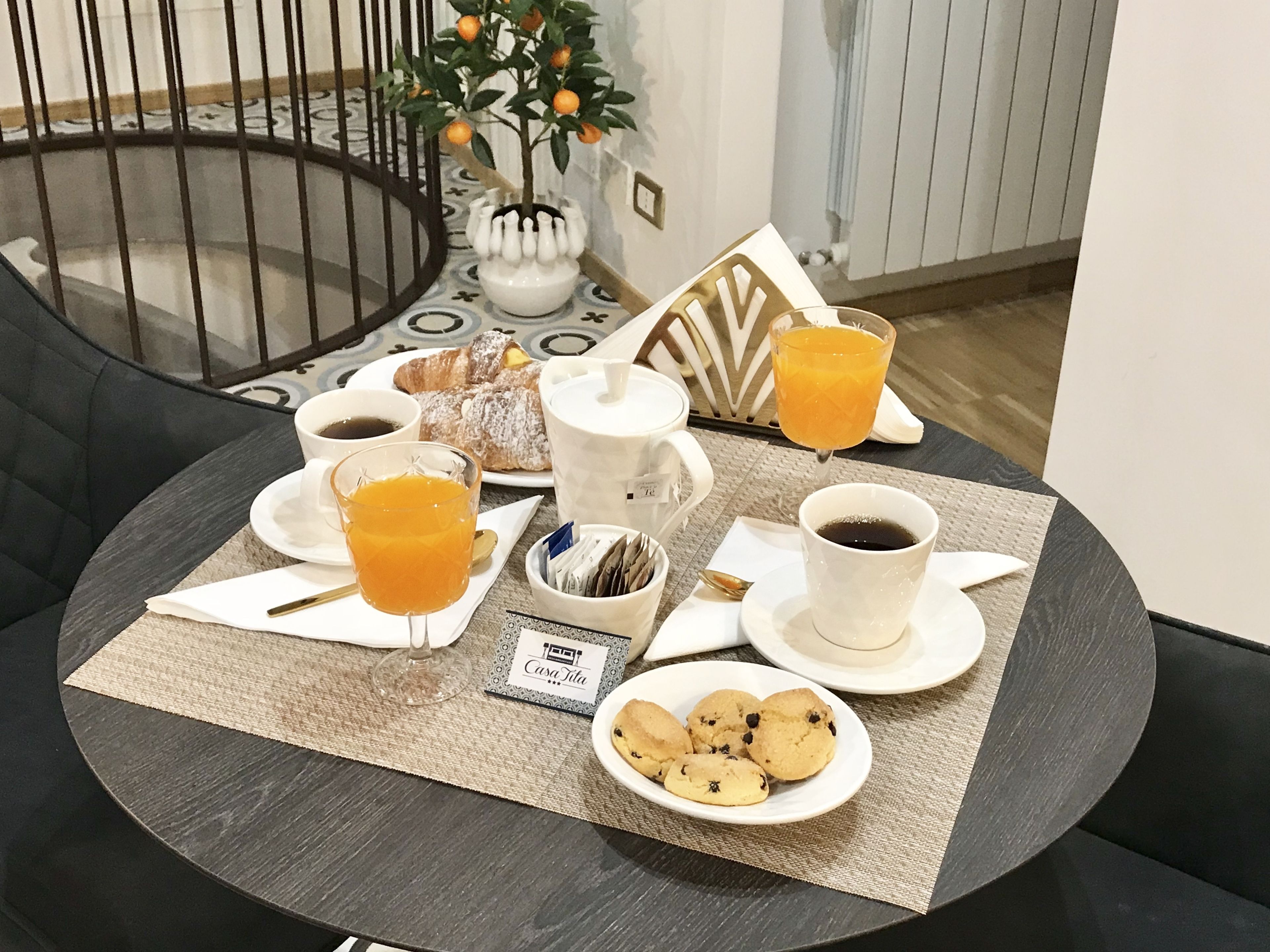 Esmorzar preparat sobre una taula rodona amb un suport, incloent croissants, suc d'aranja, cafè i galetes.