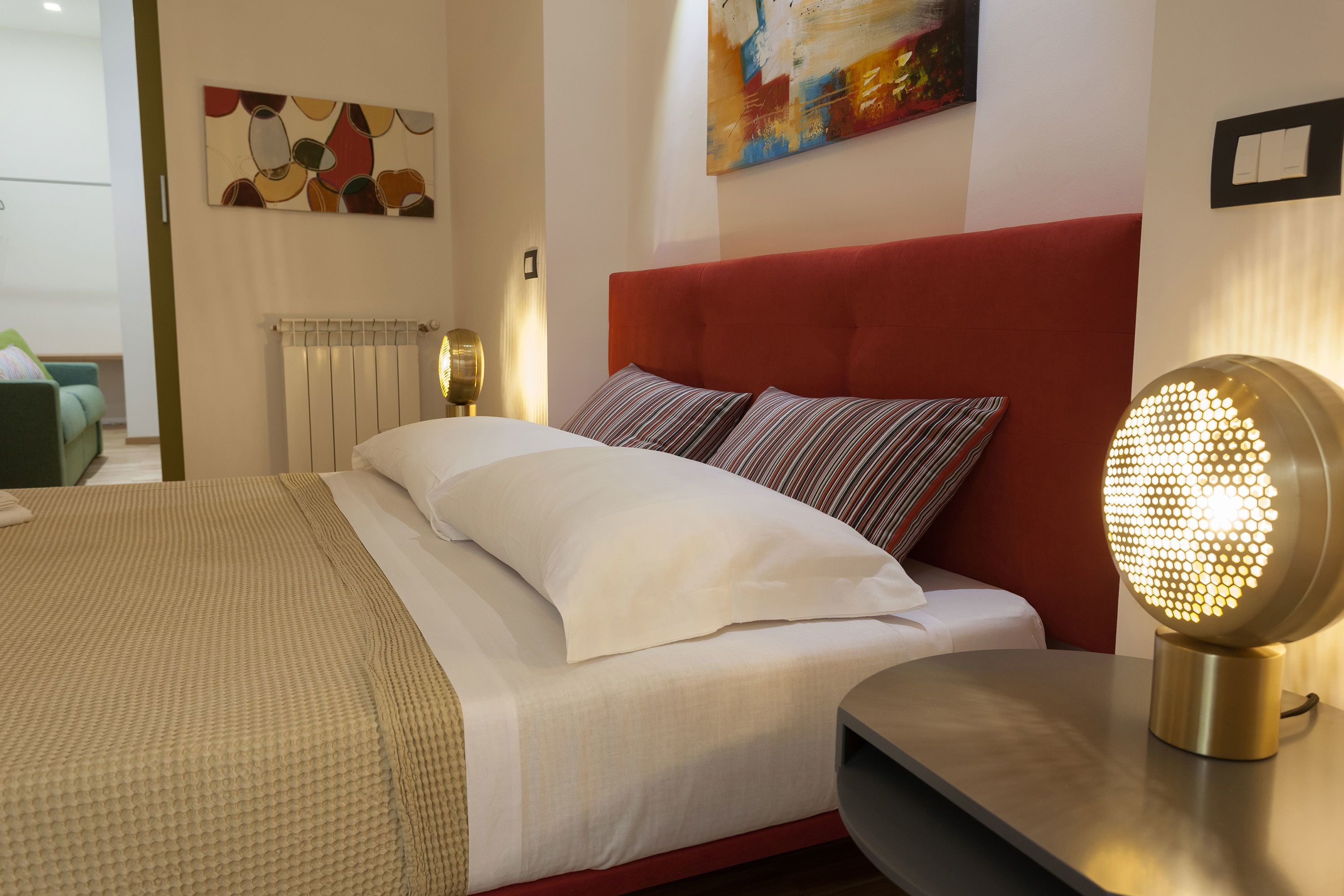 A neatly made bed with white linens, a red upholstered headboard, and a bedside table with a round, illuminated lamp.