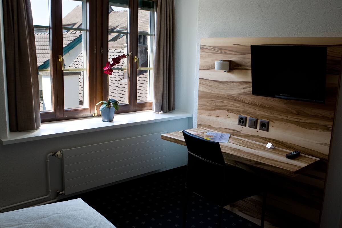 Ein Hotelzimmer mit einem Holztisch, einem an der Wand befestigten Fernseher, einem Fenster mit Vorhängen und einer Heizung.