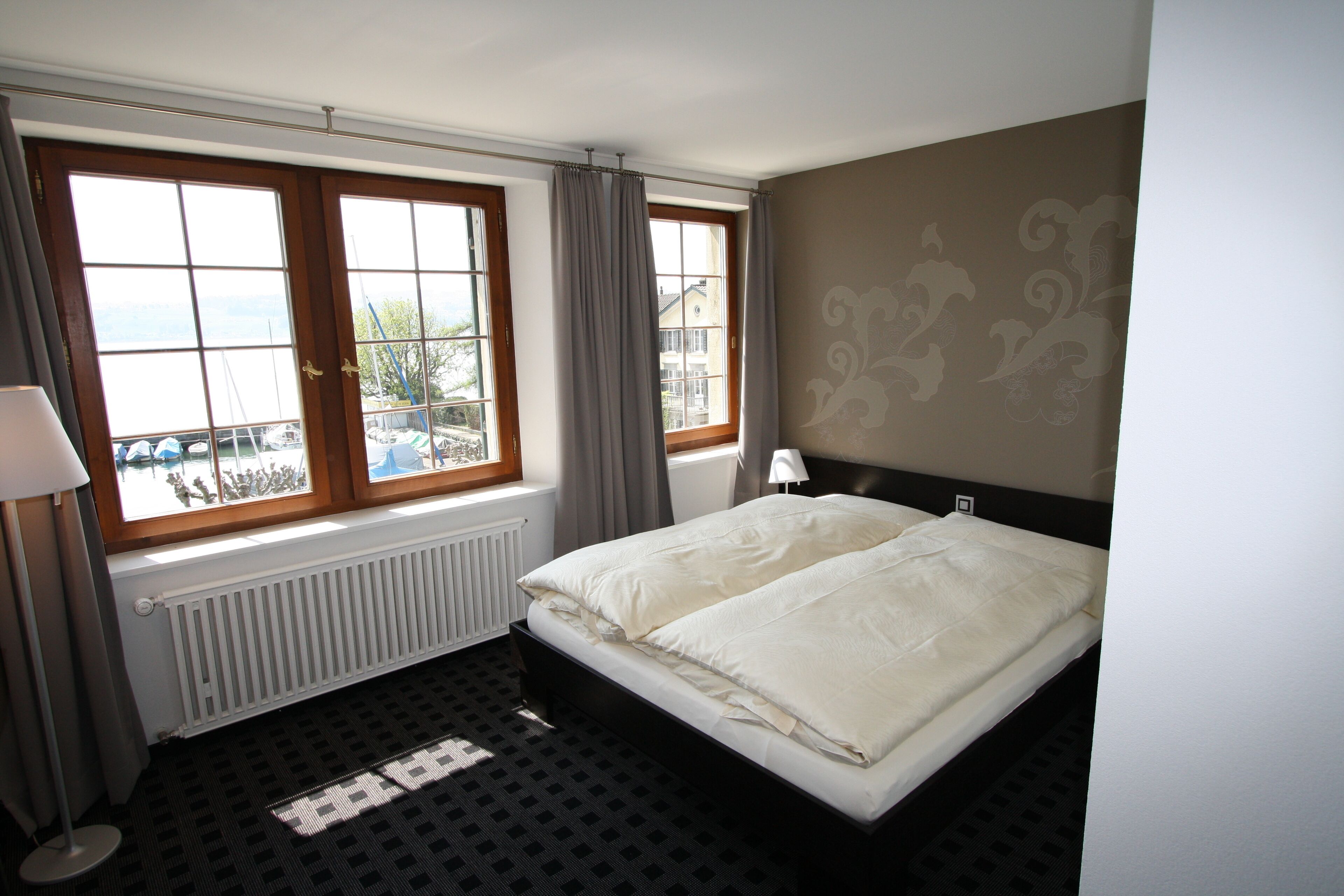 A hotel room with a bed, a window with curtains, a radiator, and a view of a body of water.