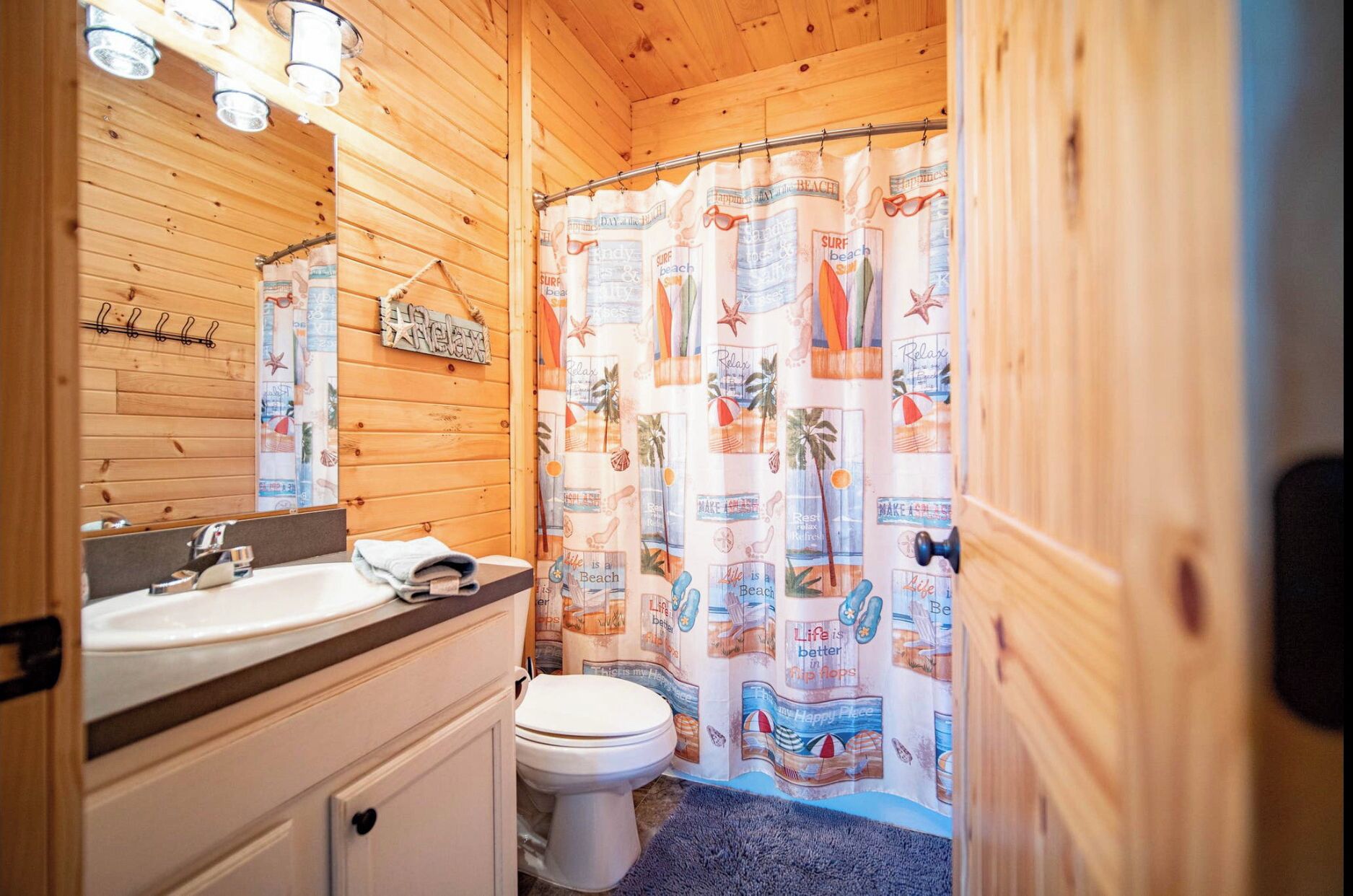 A wooden cabin bathroom with a wooden vanity, a toilet, and a shower curtain with beach-themed designs.
