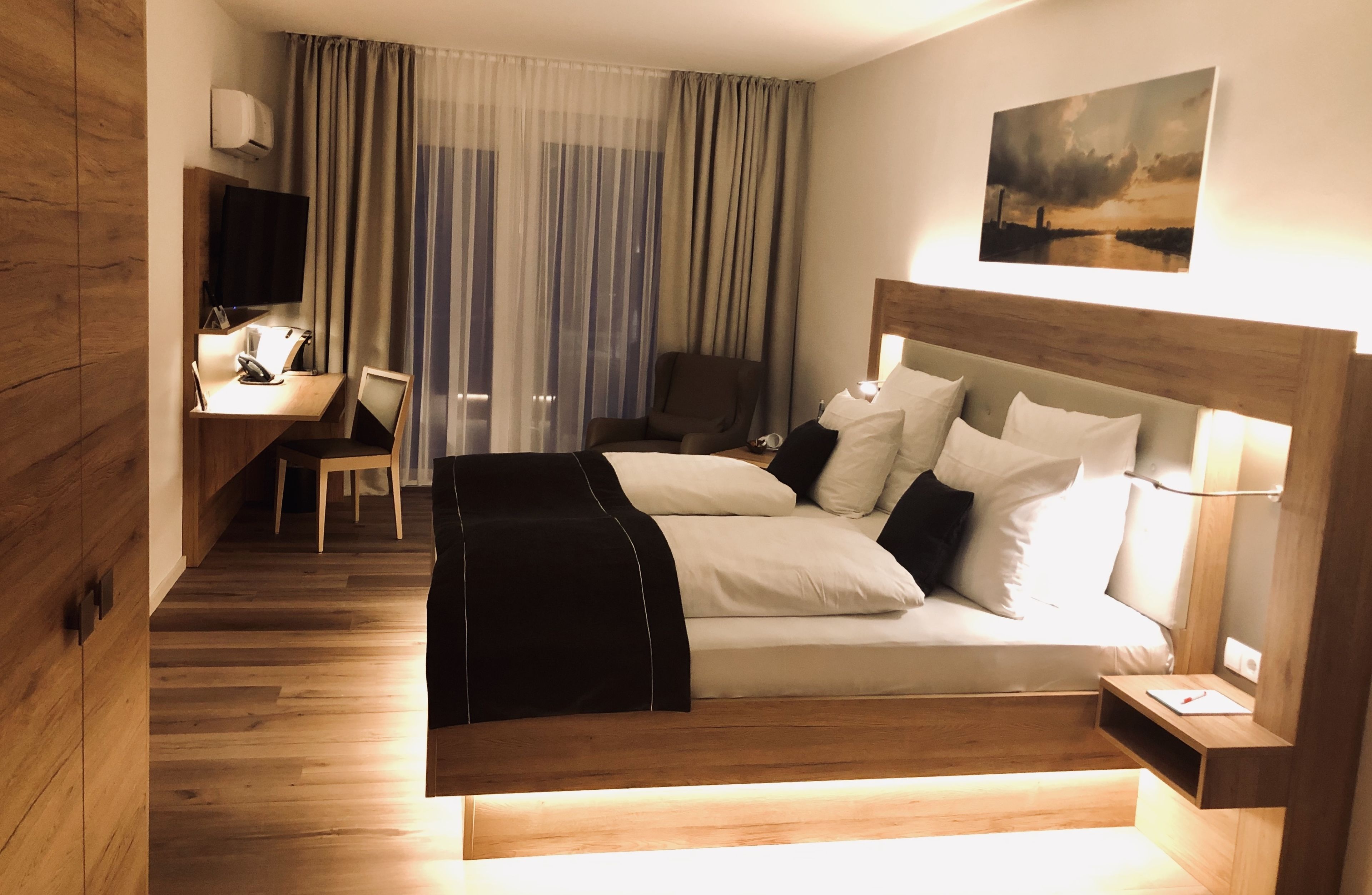 A modern hotel room with a wooden floor, a bed with a headboard, a desk with a chair, a TV, and a large window with curtains.