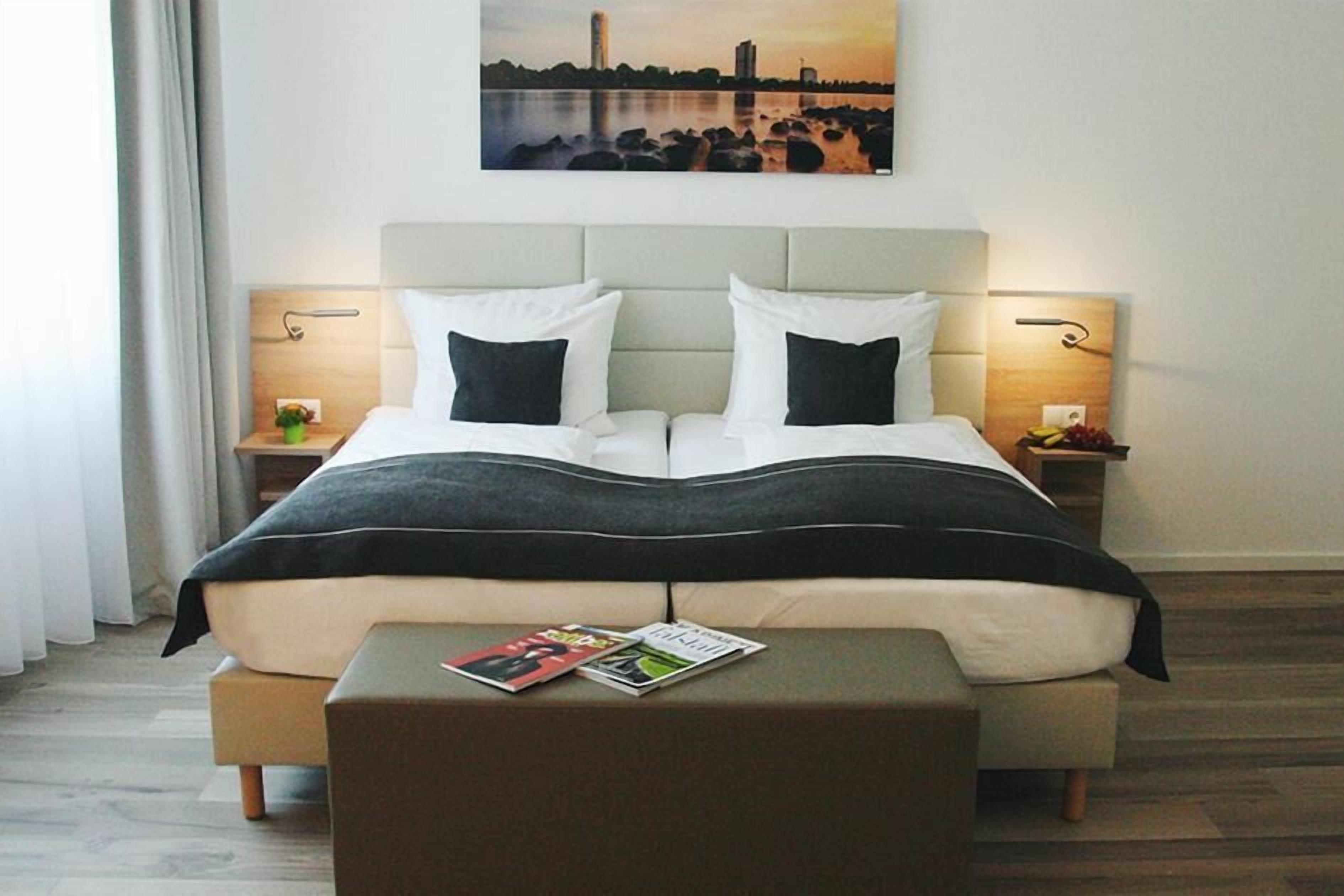 Quarto de hotel com uma cama grande, duas mesas de cabeceira, um criado-mudo, um banco e uma pintura com vista para a cidade na parede.