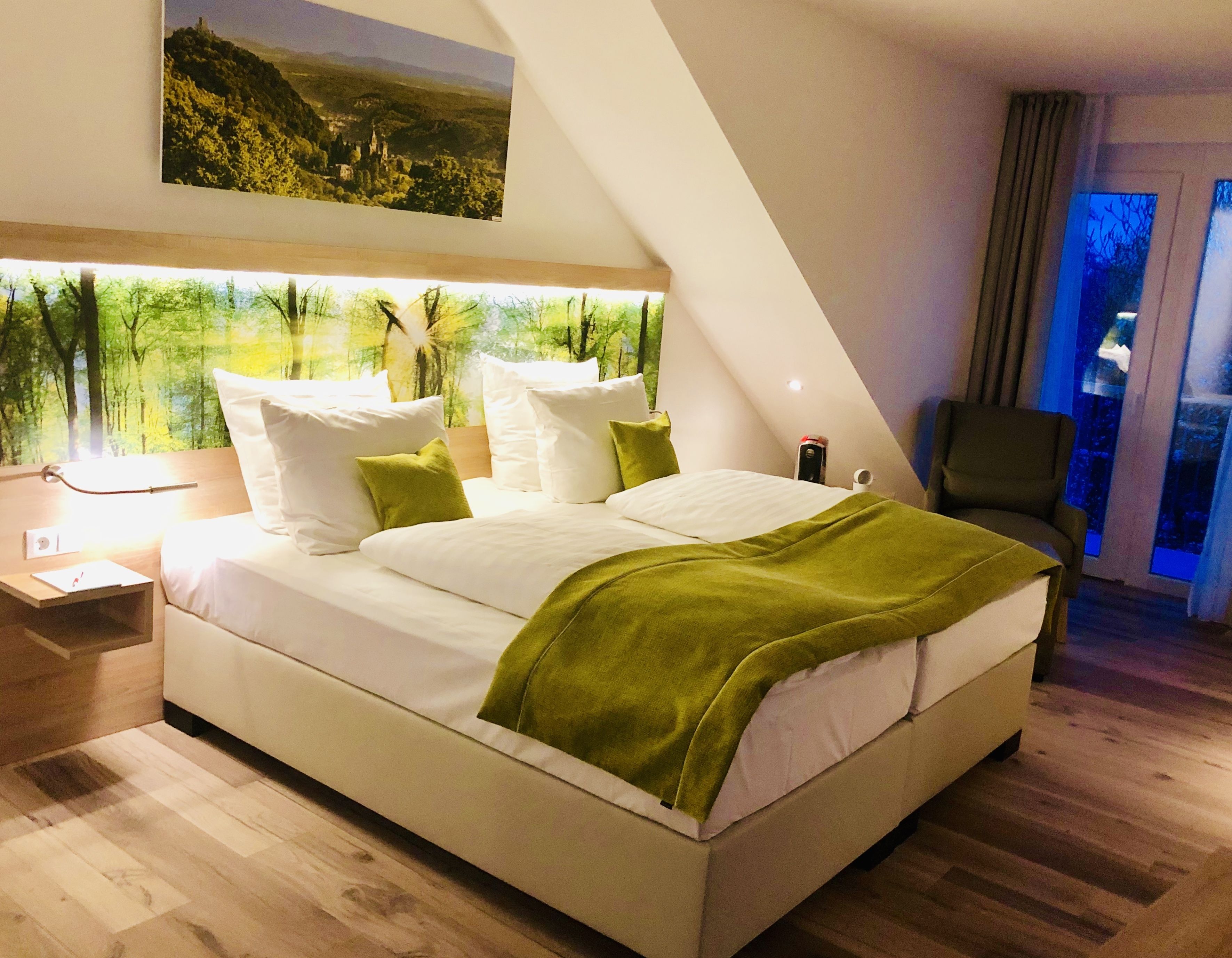 A modern hotel room with a large bed, a nightstand, a chair, and a painting of a forest on the wall.