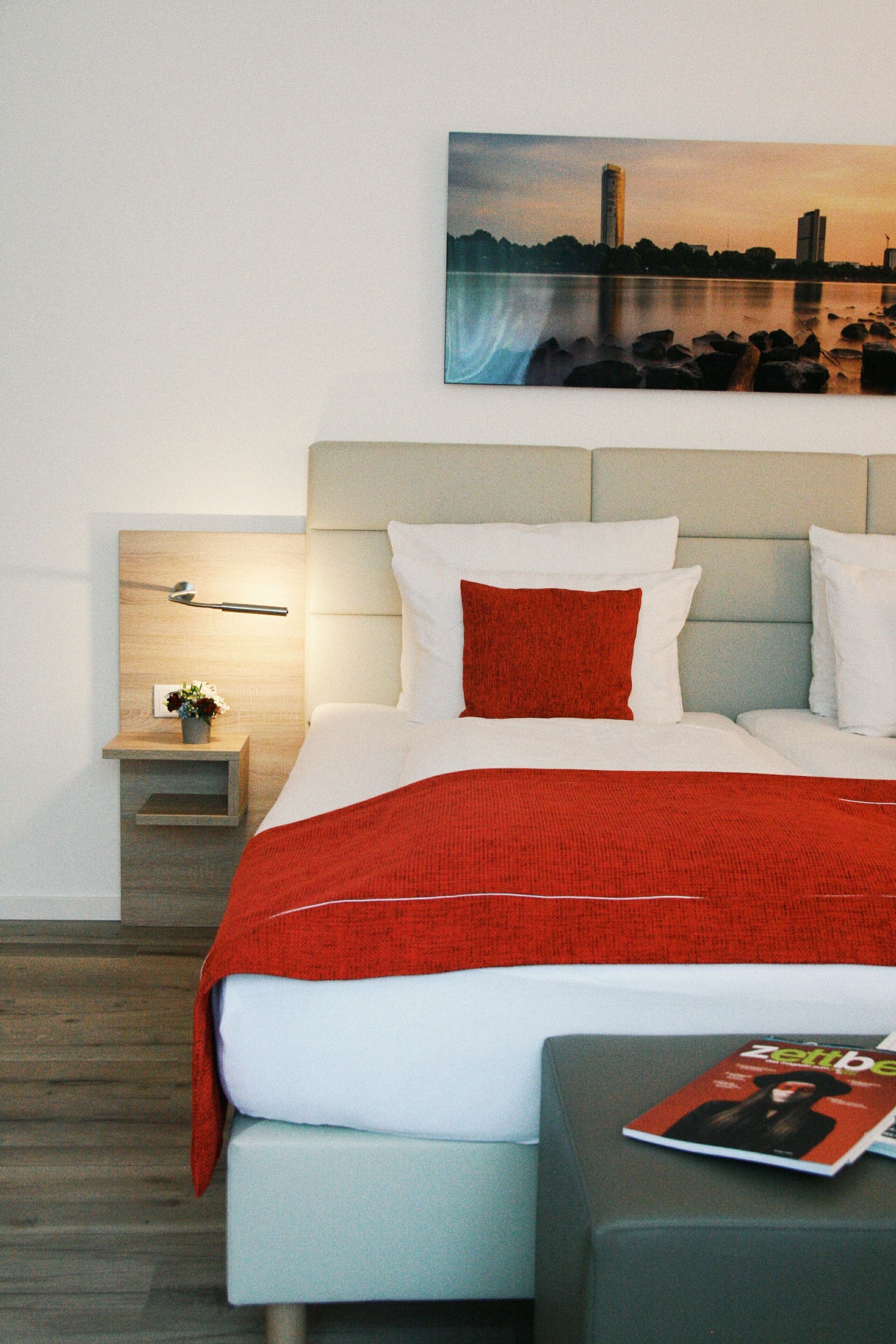 Una habitación de hotel con una cama, una mesita de noche, una revista y un cuadro en la pared.