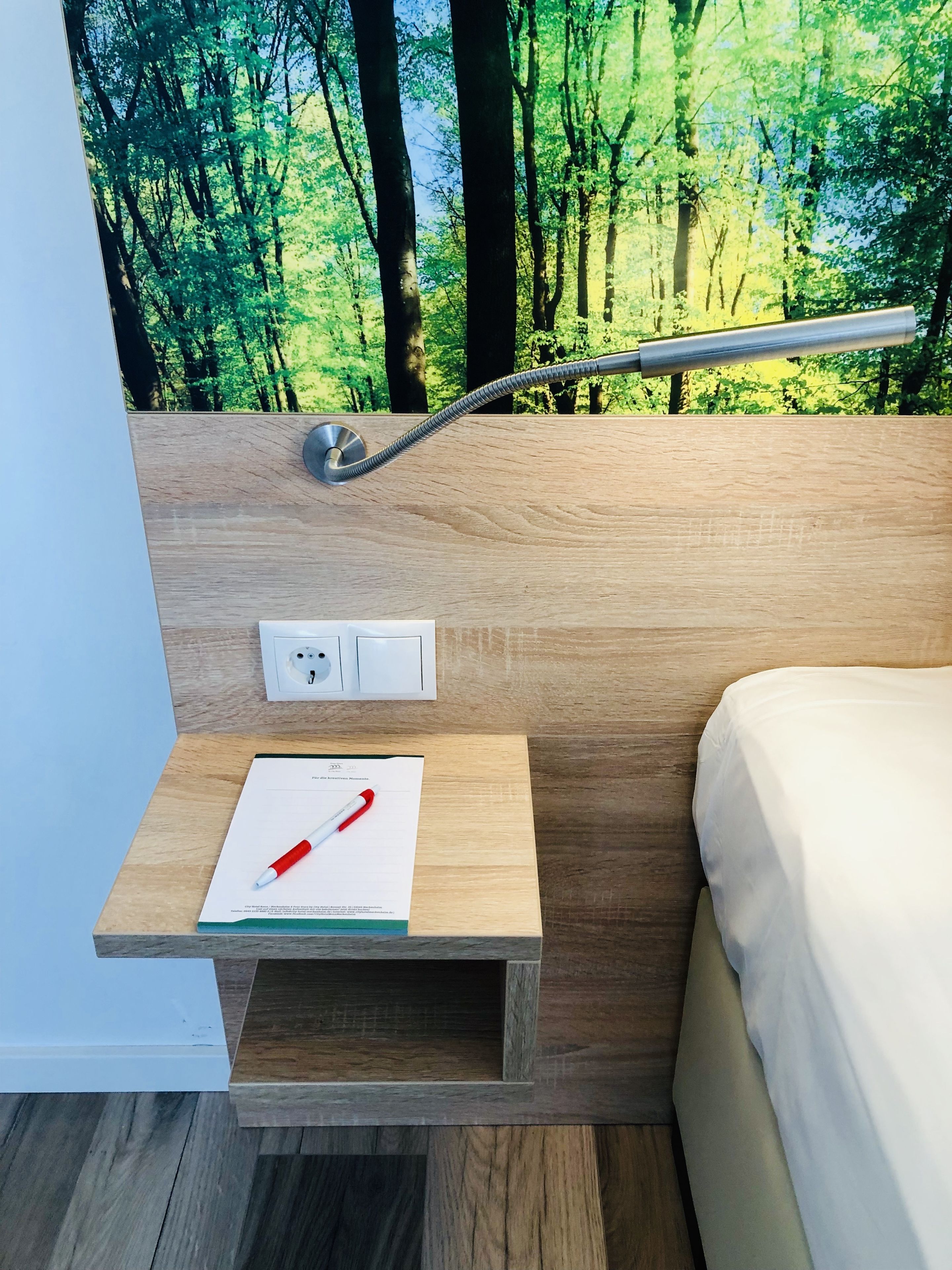 Un dormitorio moderno con cabecera de madera, una mesita de noche con lámpara y vistas a un bosque por la ventana.