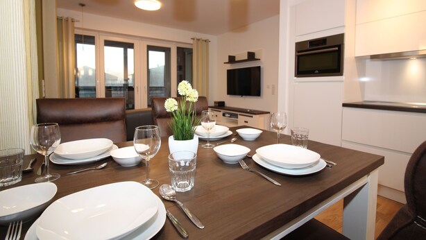 A dining table set for four with white plates, glasses, and cutlery, a vase with flowers, and a view of the city through large windows. - Wohnen am Yachthafen W45 (Cuxhaven)