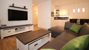 A modern living room with a flat-screen TV, a wooden coffee table, and a grey sofa with green cushions. - Wohnen am Yachthafen W45 (Cuxhaven)