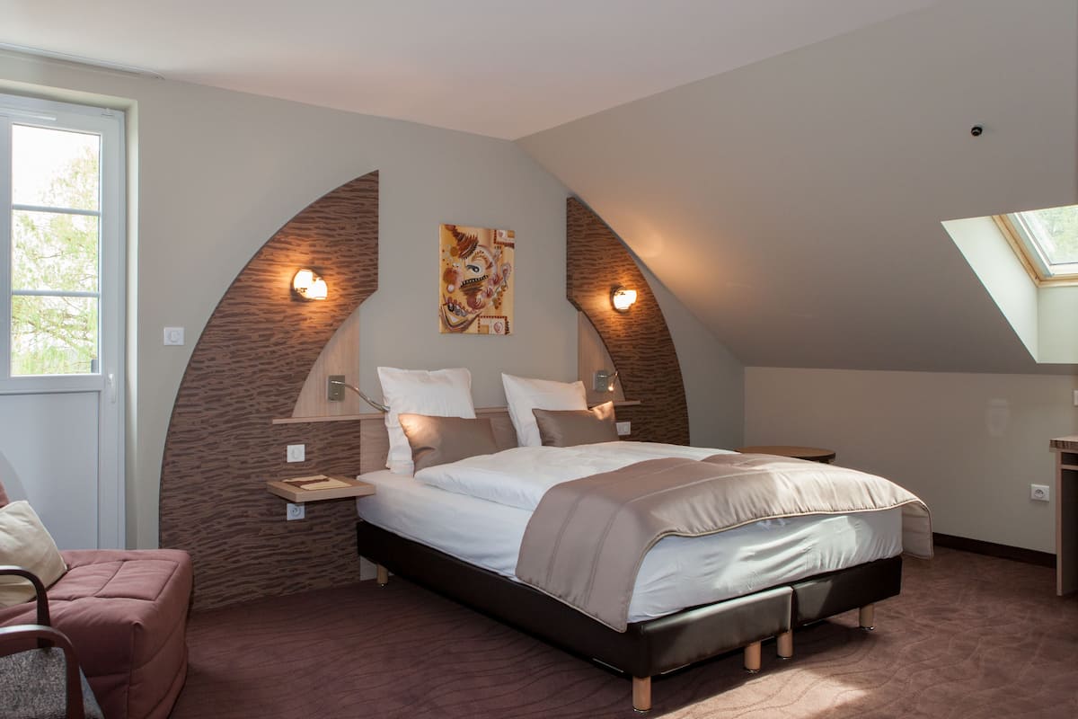 A bedroom with a large bed, a chair, a nightstand, and a wall-mounted lamp.