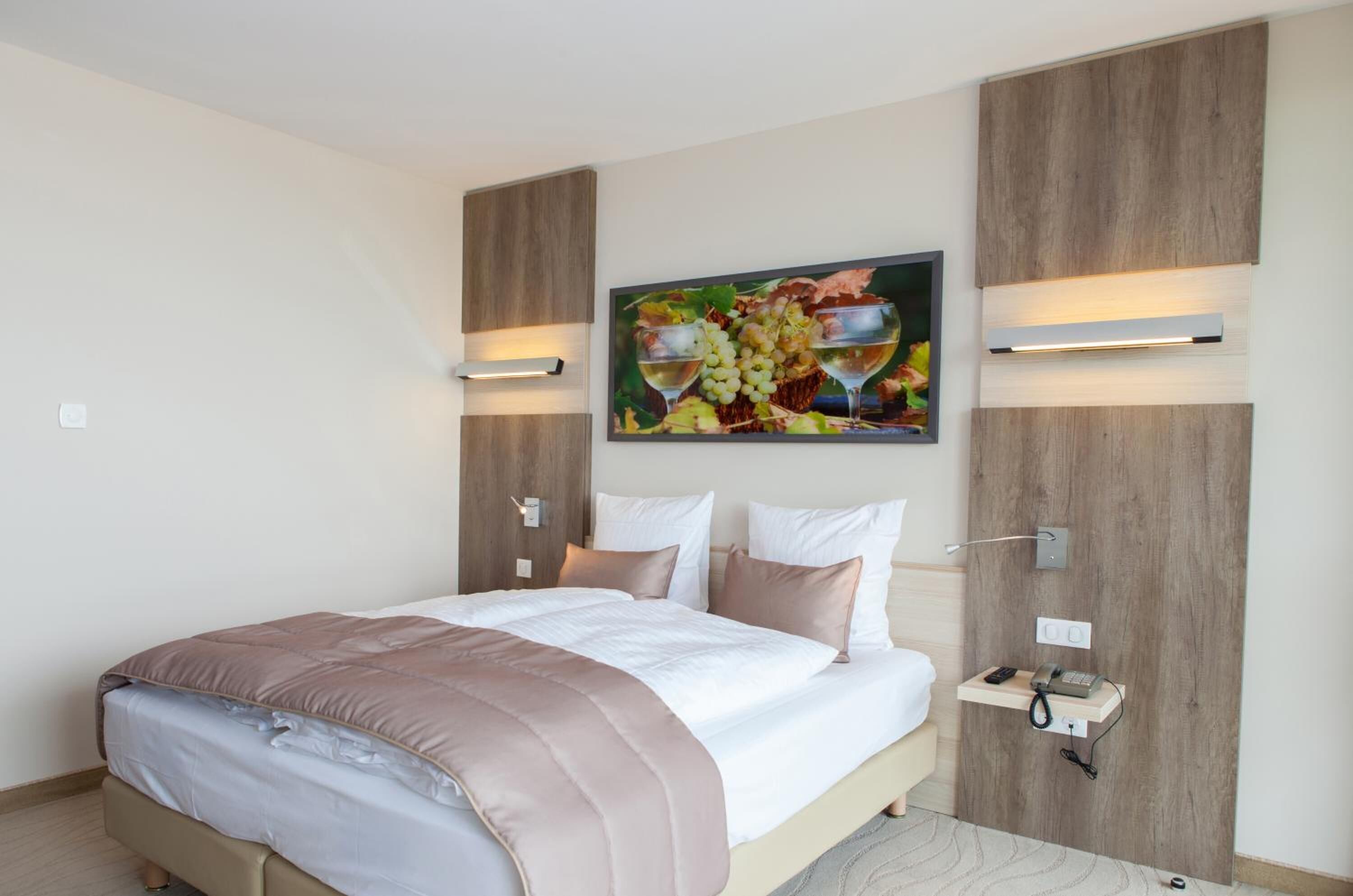 A modern hotel room with a large bed, a bedside table with a phone and a lamp, and a wall-mounted artwork of grapes and wine glasses.