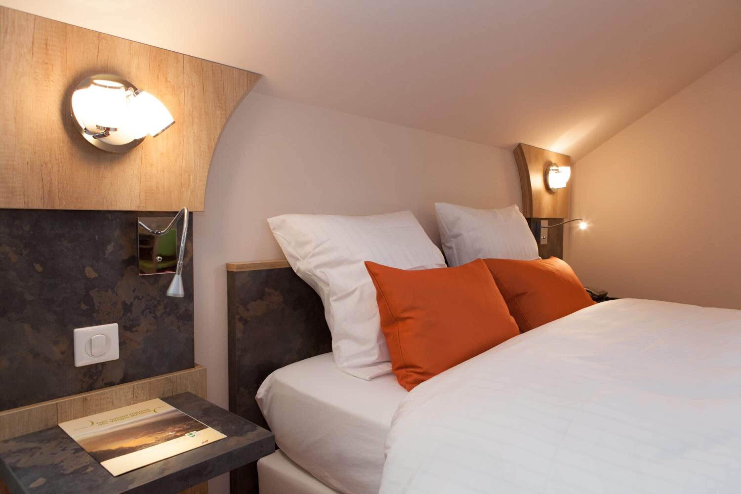 Een hotelkamer met een bed, twee kussens, een nachtkastje, een lamp en een wandlamp.
