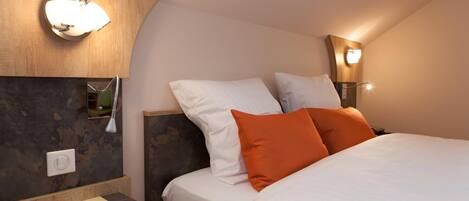 Habitación de hotel con cama, dos almohadas, mesita de noche, lámpara y aplique de pared.