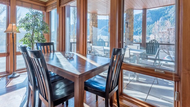 A dining area with a wooden table and chairs, large windows with wooden frames, and a view of a mountainous landscape.