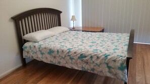 A single bed with a floral bedspread, a wooden headboard, a bedside table with a lamp, and a window with blinds.