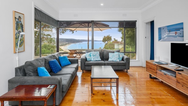 A spacious living room with a large window offering a view of the sea, a grey sofa set, a wooden coffee table, and a TV on a wooden stand.