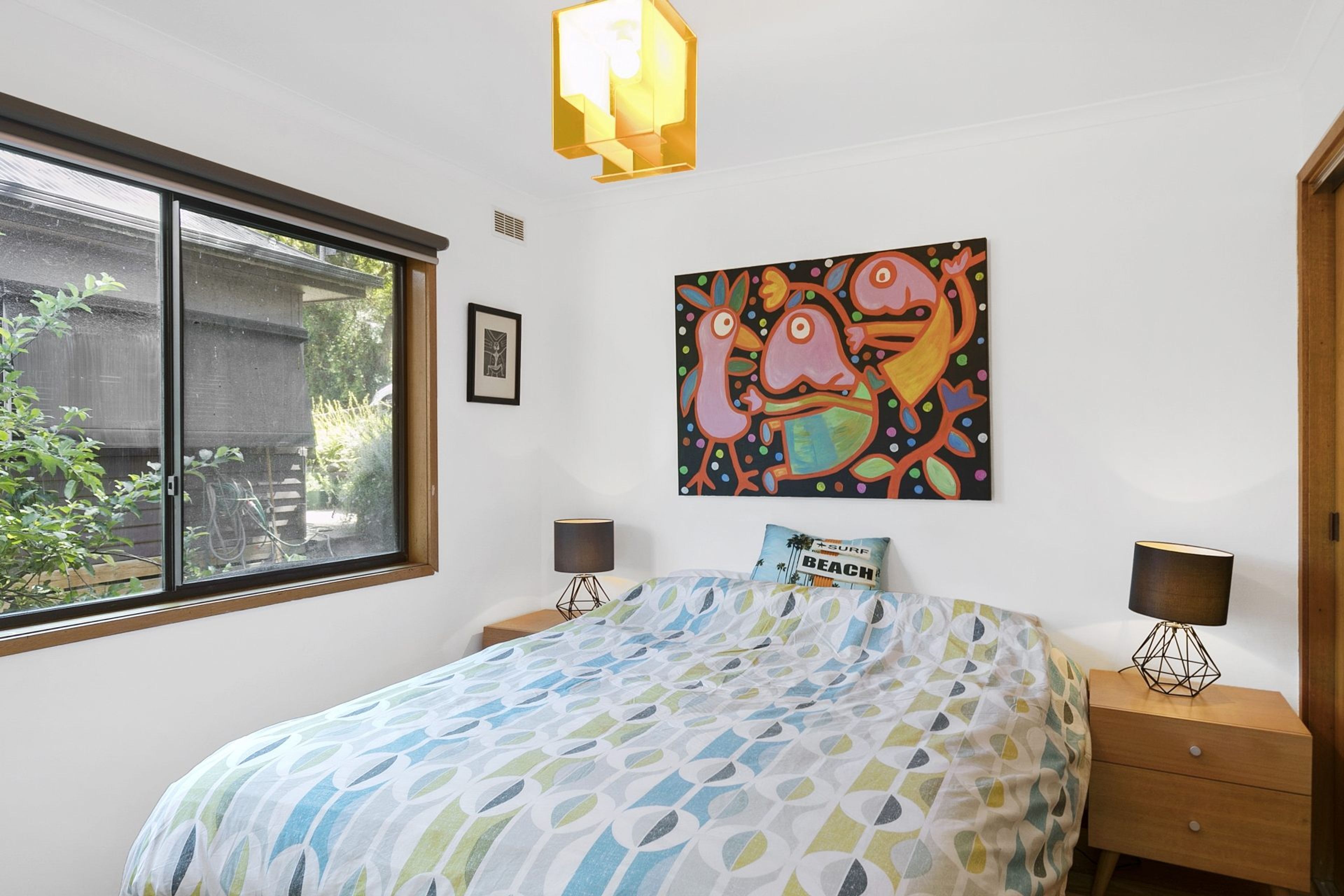 A bedroom with a bed, two bedside tables, a window, a wall-mounted painting, and a door.