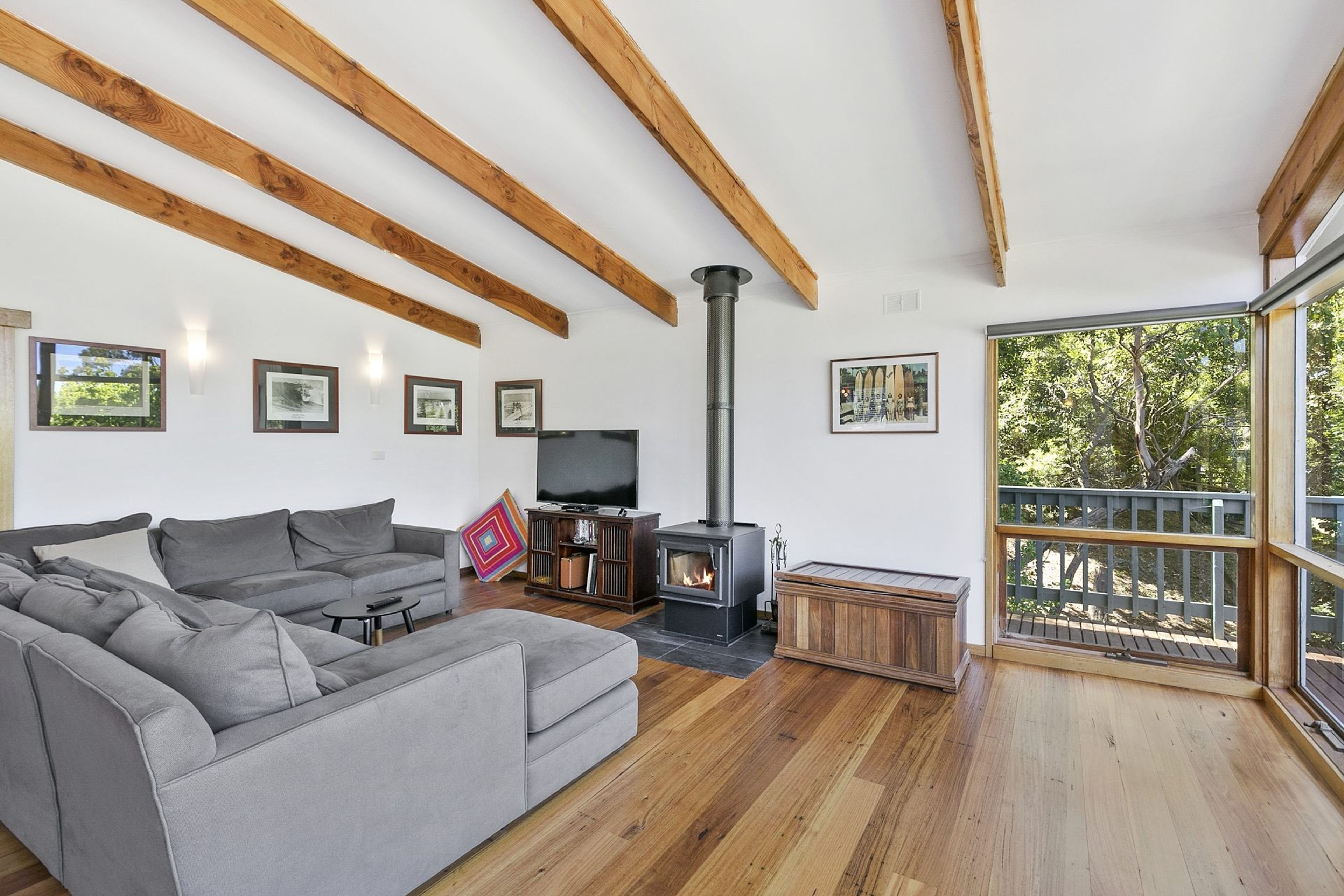 A modern living room with a grey sofa, wooden beams, a fireplace, and a balcony with a view of trees.