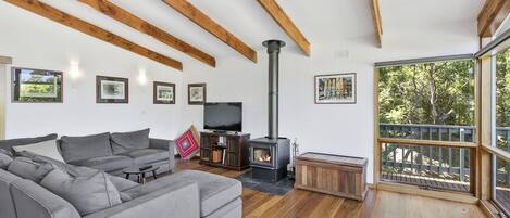 A modern living room with a grey sofa, wooden beams, a fireplace, and a balcony with a view of trees.