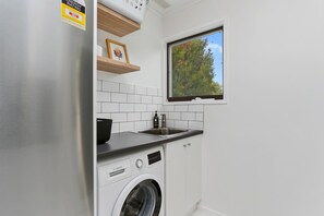 A compact kitchen with a built-in washing machine, a stainless steel refrigerator, and a window with a view of trees. - The Blue House (Apollo Bay)
