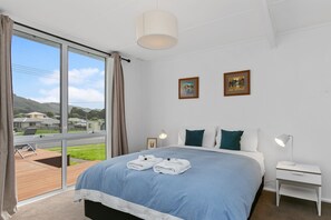 A bedroom with a large window, a bed with blue bedding, two pillows, a bedside table with a lamp, and two framed artworks on the wall. - The Blue House (Apollo Bay)