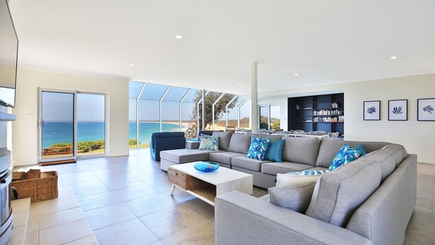 Een ruime woonkamer met een grote grijze hoekbank, een salontafel en een boekenkast. Er is een groot schuifglasraam dat leidt naar een balkon met uitzicht op de oceaan.