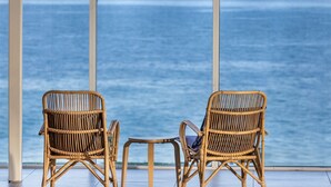 Twee rieten stoelen en een kleine tafel bij een groot raam met uitzicht op zee.