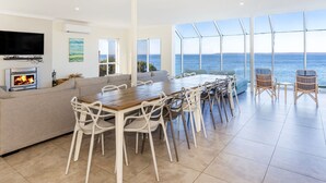 Een moderne woonkamer met een open haard, een bank, een eettafel met stoelen en een groot raam met uitzicht op zee.