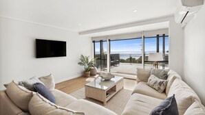 A modern living room with a flat-screen TV, a sofa set, a coffee table, and a view of the outdoors through large windows. - Apartment 12 Pacific Apartments (Lorne)