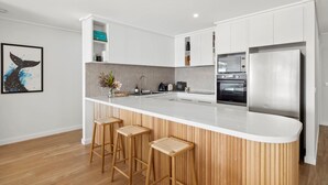 A modern kitchen with a white countertop, wooden stools, and stainless steel appliances. - Apartment 12 Pacific Apartments (Lorne)