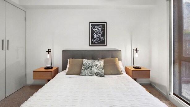 A bedroom with a bed, two bedside tables, a wall-mounted light, and a window. - Apartment 12 Pacific Apartments (Lorne)
